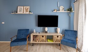 A living room with a blue wall, a flat-screen TV, two blue armchairs, a wooden coffee table, and shelves with decorative items.