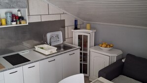 A compact kitchenette with a built-in stove, a sink, and a small countertop. There is a white cabinet with glass doors, a white chair, and a small table with a bowl of fruit. - Apartment Jadranka - Two Bedroom Apartment With Balcony And Sea View (Korcula)