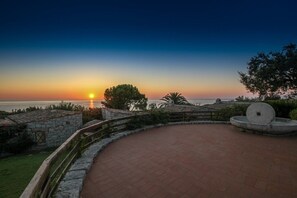 A sunset view from a paved terrace with stone walls, a circular stone feature, and a distant body of water.