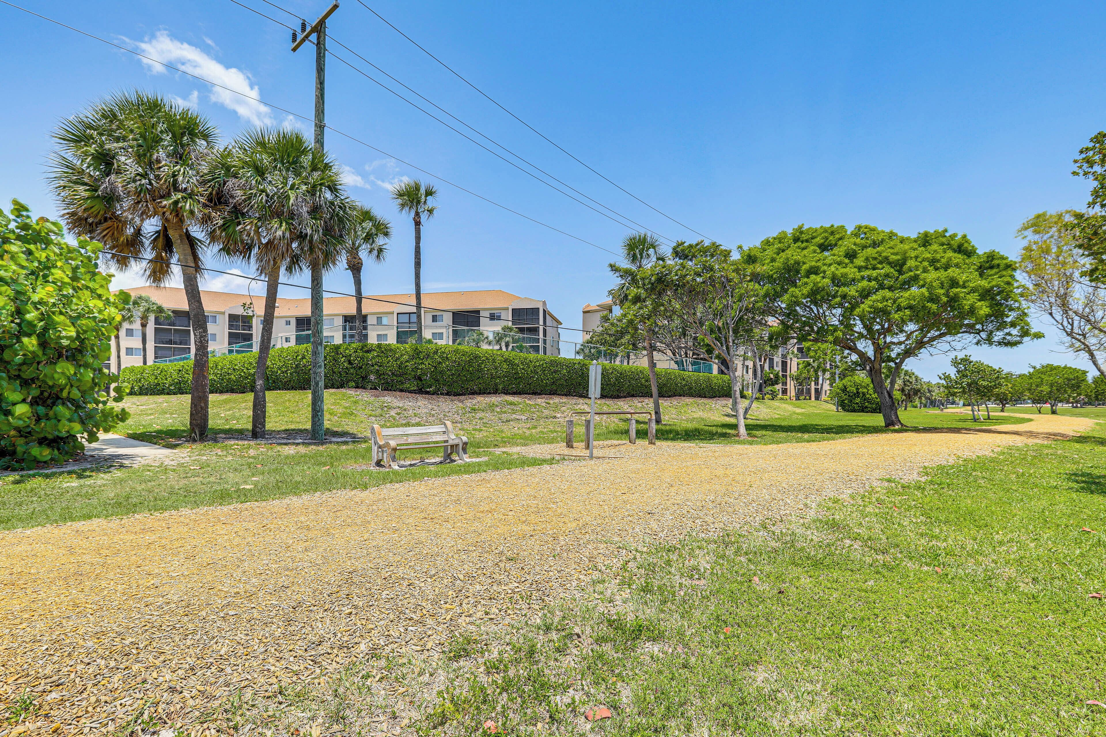 A pathway leading to a building with a flat roof, surrounded by palm trees and a grassy area with a bench.