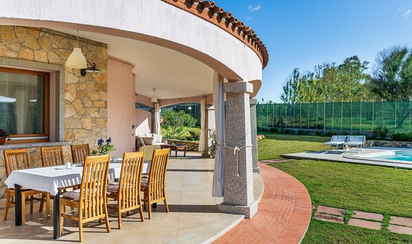 A covered outdoor dining area with a stone wall, wooden chairs, and a table.