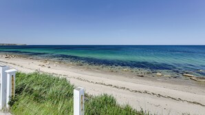 Ein Strand mit klarem, blauem Wasser, sandigem Ufer und einem weißen Zaun im Vordergrund.