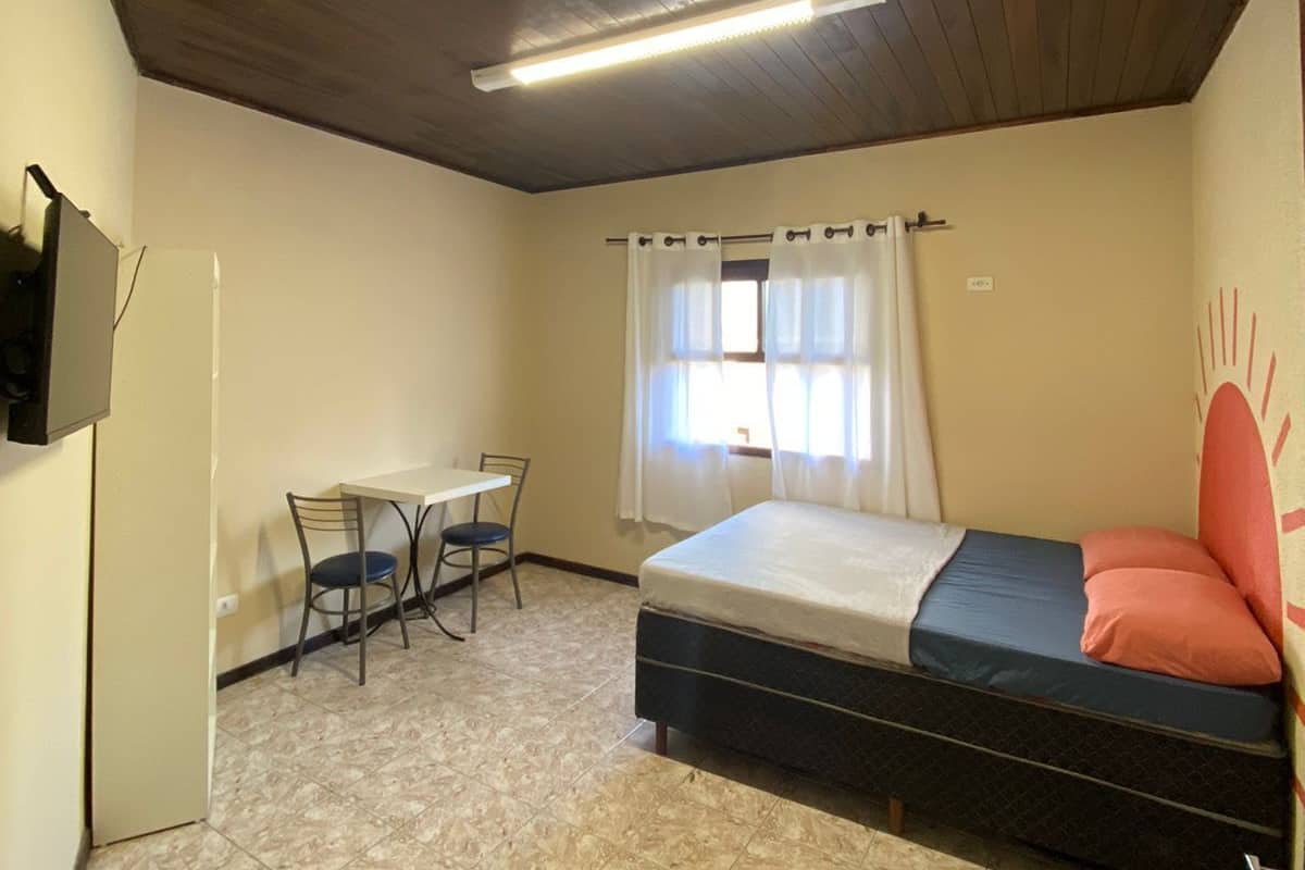 A small, single-room accommodation with a bed, a table with chairs, a window with curtains, and a ceiling light.