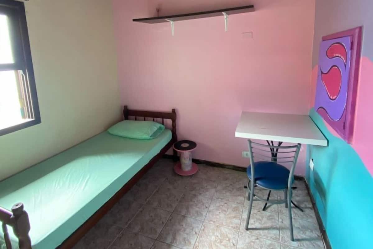 A small, single-bed room with a wooden ceiling, a window, a desk with a chair, and a pink wall with a heart-shaped decoration.