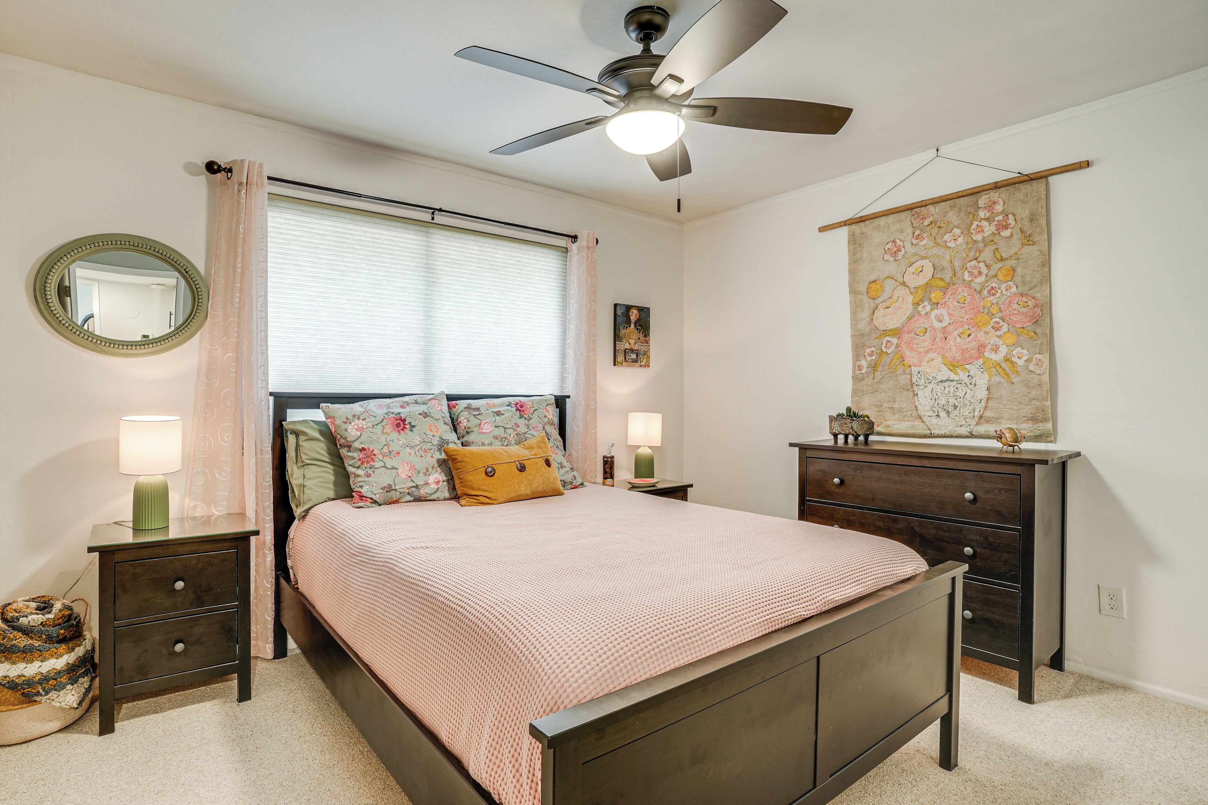 A bedroom with a bed, nightstands, a dresser, a ceiling fan, a window with curtains, a mirror, and a wall hanging.