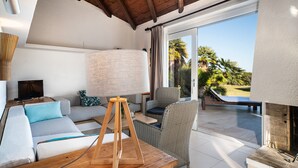Uma sala de estar moderna com um sofá, uma mesa com abajur, uma cadeira de vime e vista para o pátio externo com piscina e palmeiras.