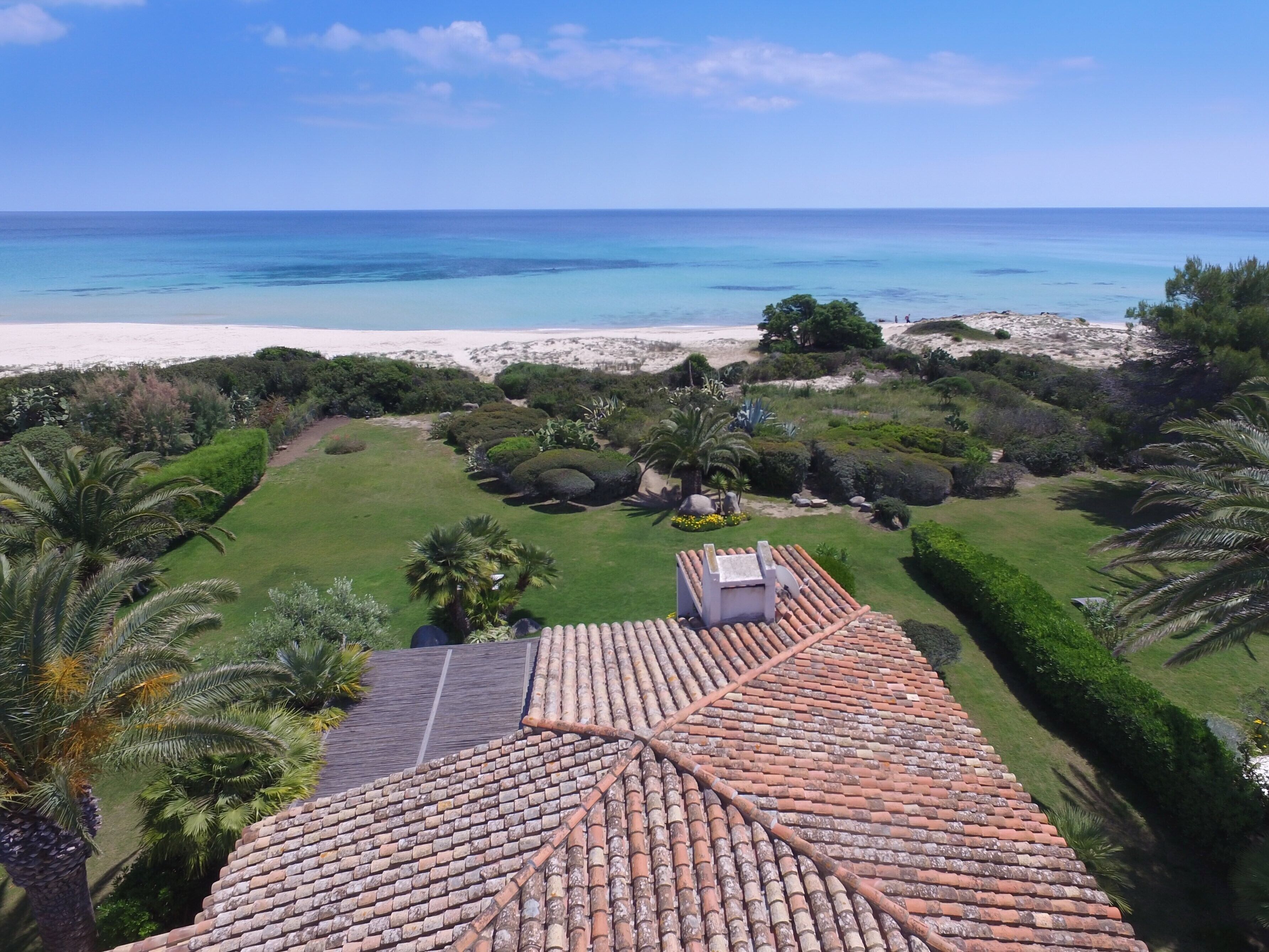 A rooftop with terracotta tiles, a well-maintained garden with various shrubs and palm trees, and a view of the beach and ocean.