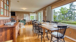 A dining area with a wooden table and chairs, a brick fireplace, and large windows with a view of trees. - Maxwelton Aerie Bluffside Retreat w/ Sweeping Puget Sound Views + Firepit (Clinton)