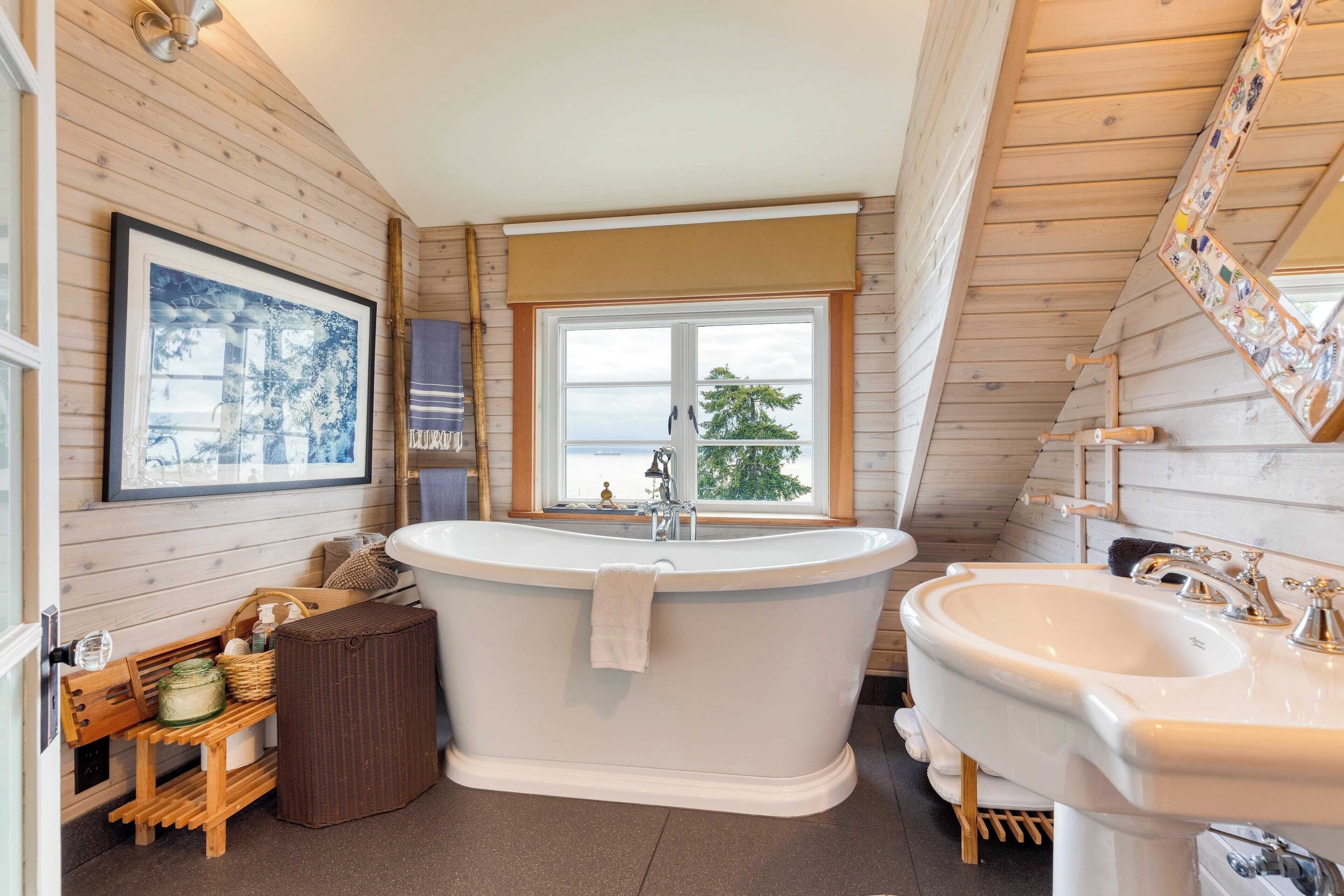 A bathroom with a freestanding bathtub, a sink, wooden walls, a window with a view, and a framed picture on the wall.