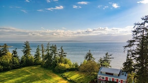 A house with a red roof and white walls, surrounded by a well-maintained lawn and tall trees, overlooking a calm body of water. - Maxwelton Aerie Bluffside Retreat w/ Sweeping Puget Sound Views + Firepit (Clinton)