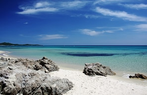 A beach with clear turquoise water, rocky foreground, and distant land.