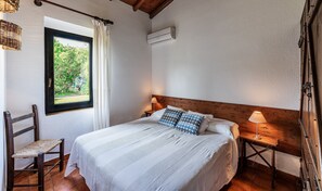 A bedroom with a bed, a chair, a window with curtains, a bedside table with a lamp, and an air conditioning unit.