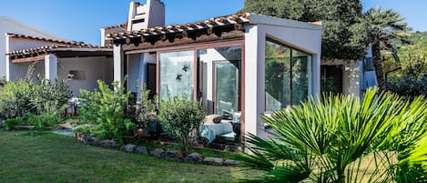 A modern house with a glass conservatory, a garden with various plants, and a clear blue sky.