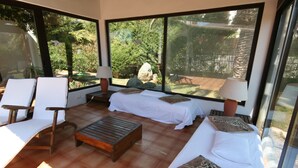 A room with a bed, two chairs, a small table, and large windows offering a view of a garden. - Villa '12' with Private Terrace, Wi-Fi and Air Conditioning (Costa Rei)