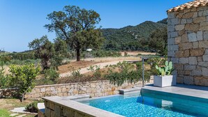 A stone pool with a clear blue pool, surrounded by a stone wall and a view of a hilly landscape with trees and a house in the distance. - Villa 'Laccus 6' with Sea View, Wi-Fi and Air Conditioning (Castiadas)