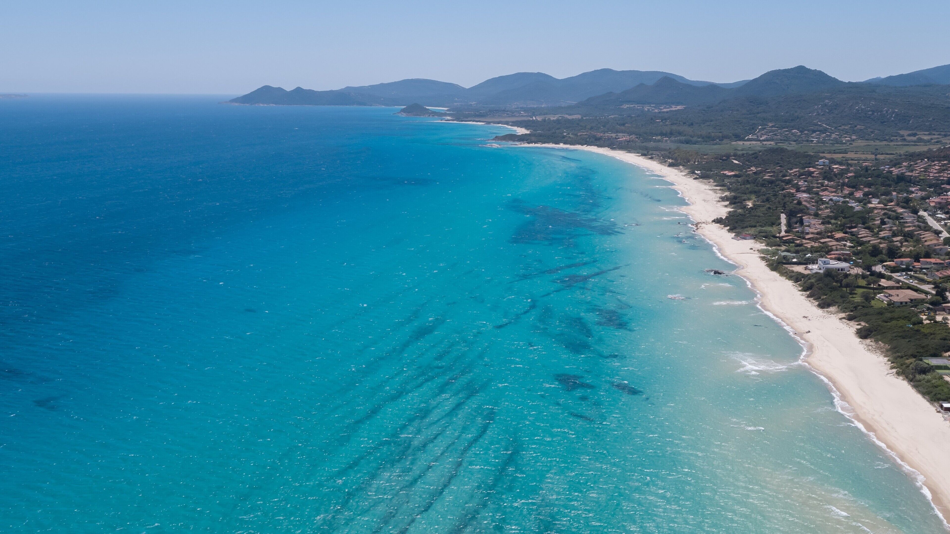 Vista aerea di una zona costiera con spiaggia, acque blu cristalline e montagne sullo sfondo.