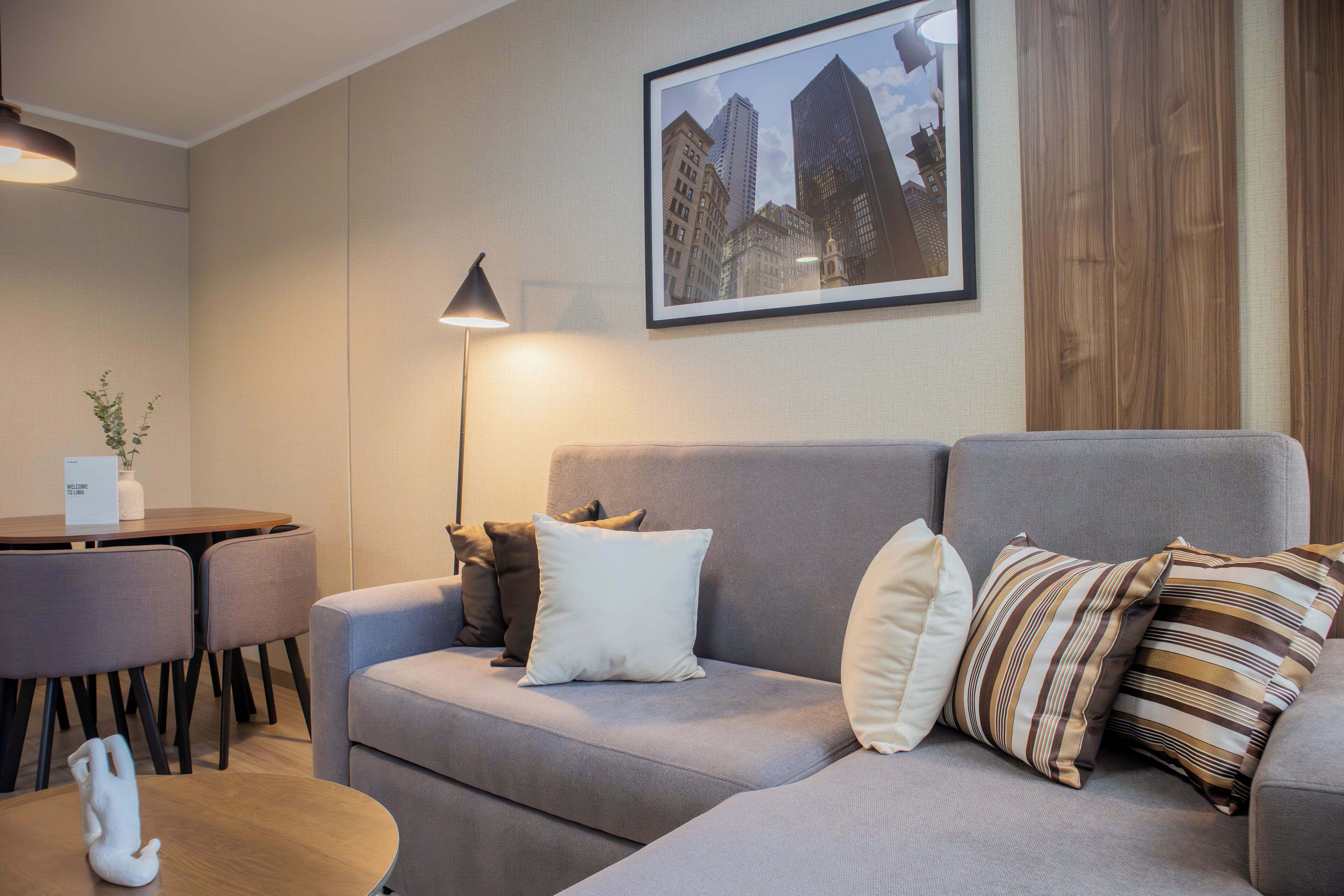 A modern living room with a grey sofa, a dining table with chairs, and a framed cityscape picture on the wall.