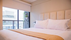 Une chambre d’hôtel moderne avec un grand lit, une vue sur la ville grâce à de grandes fenêtres du sol au plafond et une plante en pot sur le rebord de la fenêtre.