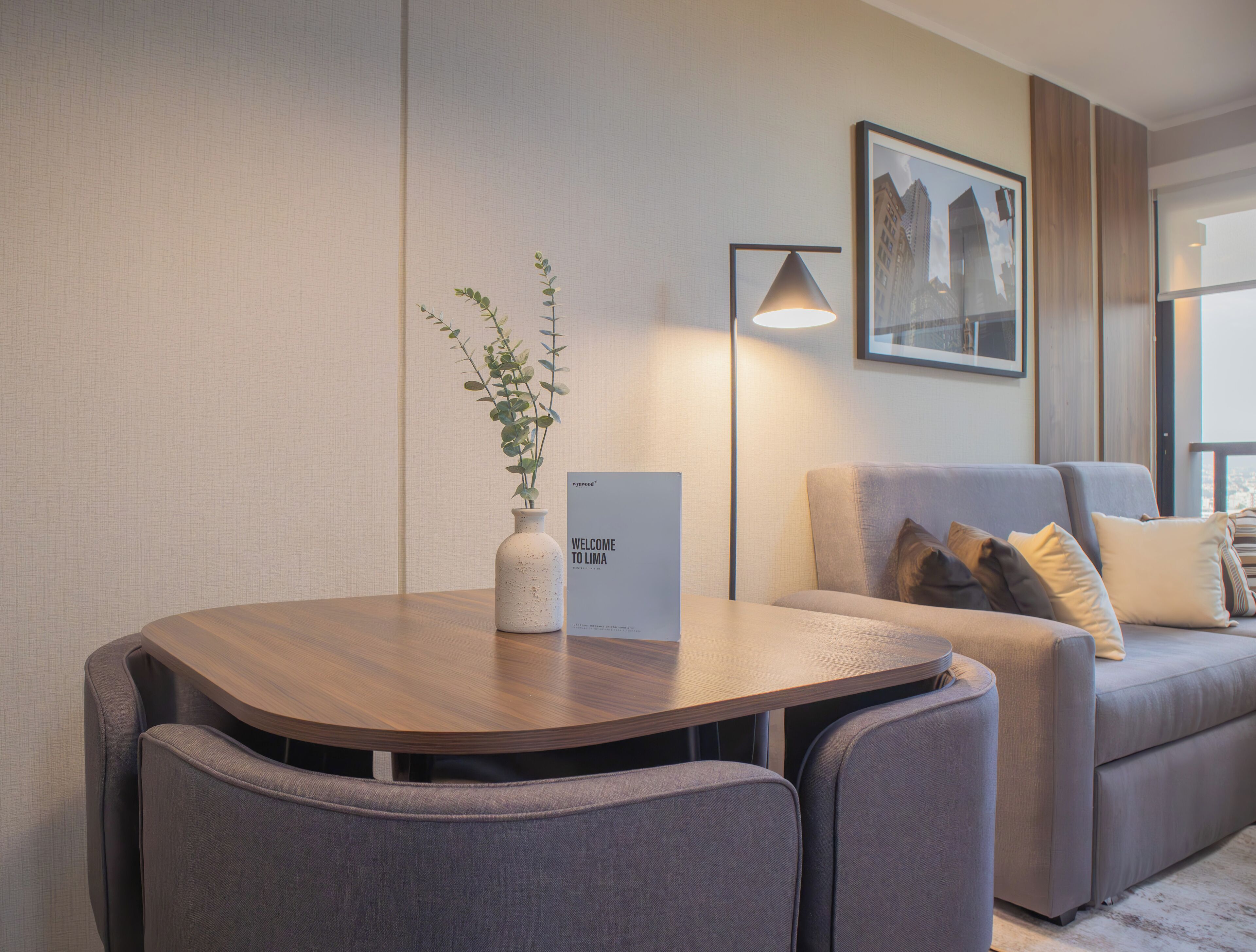 Un salón moderno con una mesa redonda de madera, dos sillones grises, un sofá gris con cojines, una lámpara de pie y un cuadro enmarcado en la pared.