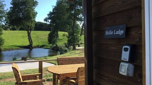 A wooden deck with a table and chairs, a sign reading "Hollie Lodge", and a view of a pond and greenery. - Hollie (Usk)