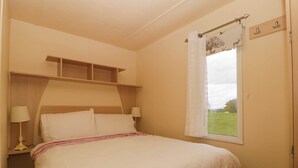 A bedroom with a bed, bedside lamps, a window with a view of a green field, and a wooden shelf above the bed. - Hollie (Usk)