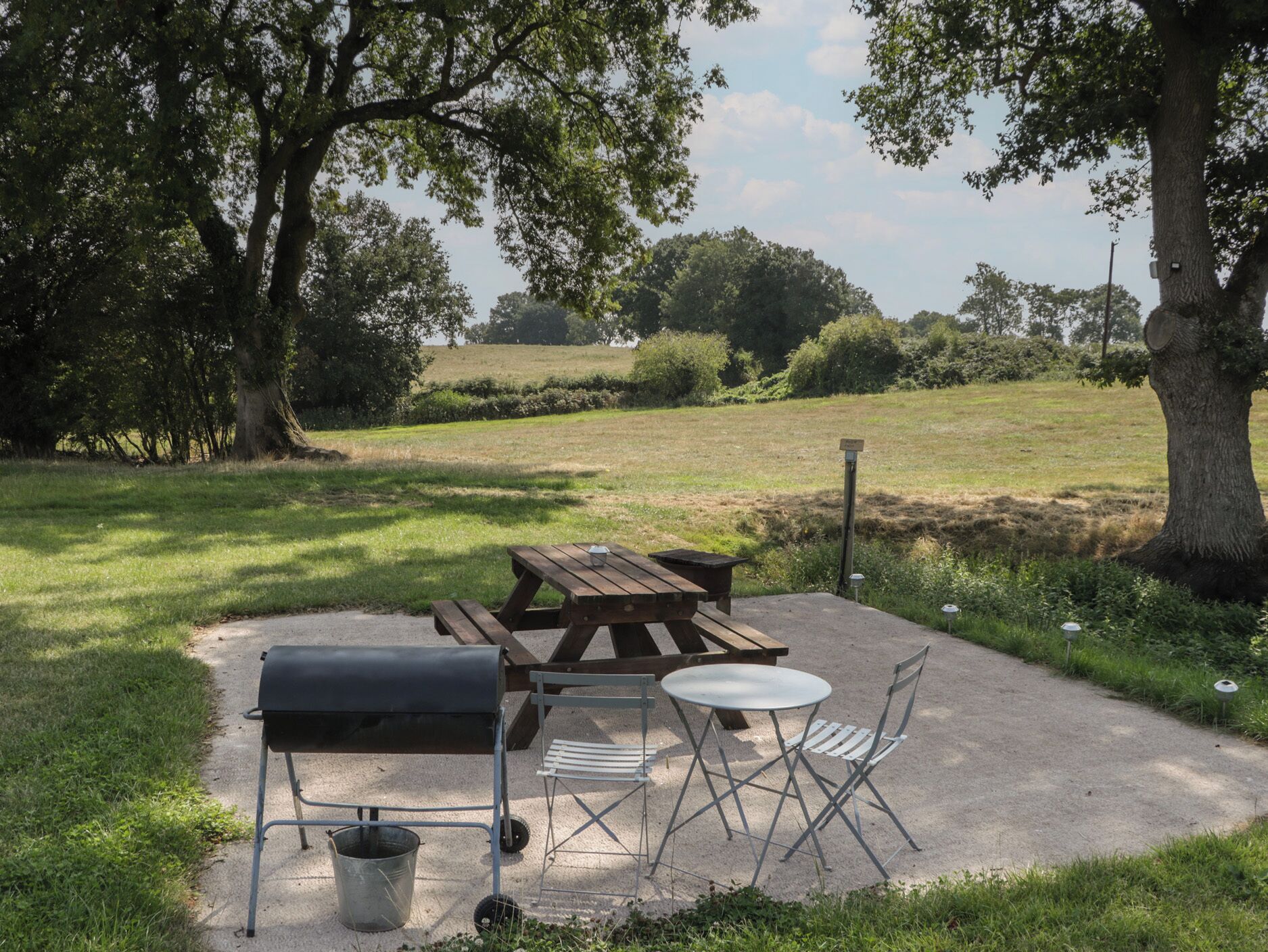 An outdoor picnic area with a table, chairs, a grill, and a trash can.