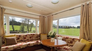 A cozy room with a patterned sofa, a round wooden table, and large windows offering a view of a green field and trees. - Hollie (Usk)