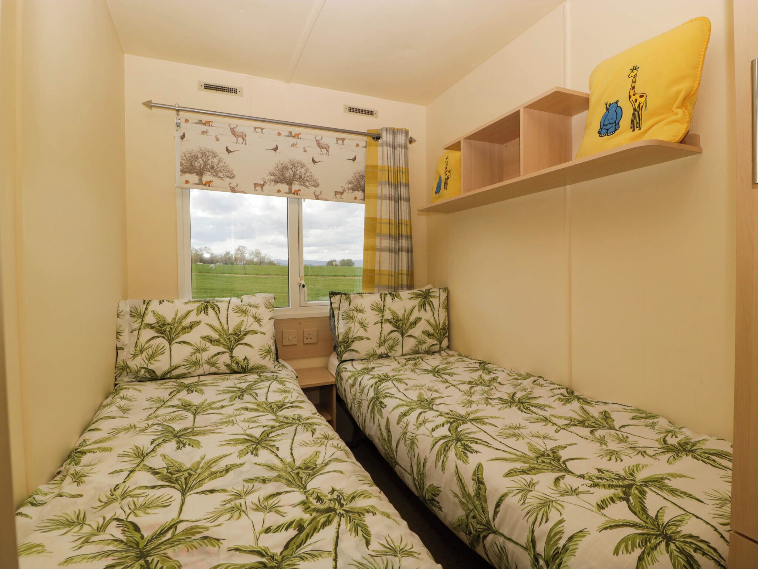 A room with two beds, a window with a view of a green field, a shelf with a yellow pillow, and a curtain with animal prints.