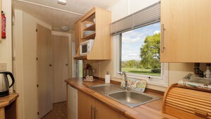 A compact kitchen with wooden cabinets, a stainless steel sink, a microwave, and a window with a view of greenery. - Hollie (Usk)