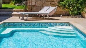 Ein Schwimmbecken mit gefliesten Rändern, Liegestühlen und einer Steinsäule im Hintergrund.