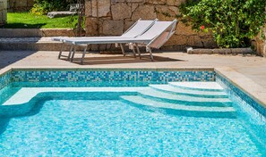 Una piscina con bordi piastrellati, sdraio e un muro in pietra sullo sfondo.