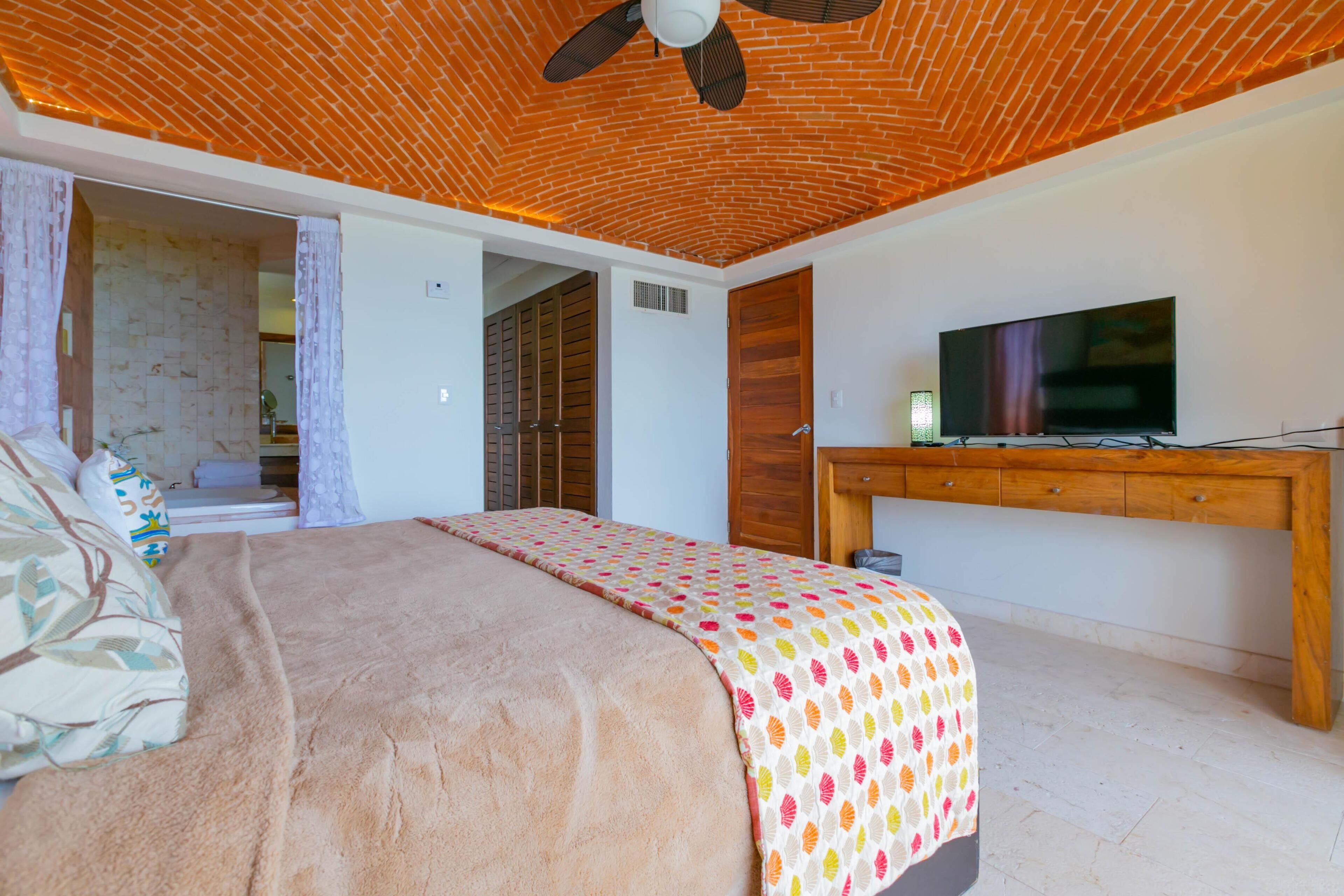 A bedroom with a bed, a TV, a wooden desk, and a ceiling fan.