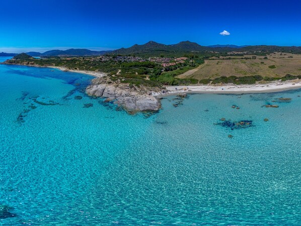 Vista aérea de una zona costera con aguas turquesas cristalinas, afloramientos rocosos y una cadena montañosa distante.