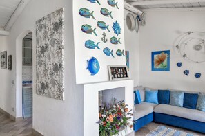 Een kamer met een witte muur, een blauwe bank, een open haard met een bloemstuk, en wanddecoratie inclusief vissen en een bloem.