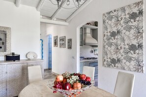 Een eethoek met een tafel voor vier personen, een fruitschaal, een witte kast met decoratieve objecten, en een keuken op de achtergrond.