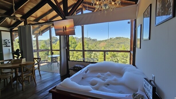 A room with a large window, a hot tub, wooden furniture, and a chandelier. - Chalés Luz do Luar (Campos do Jordao)