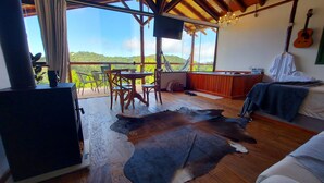 A room with a wooden floor, a dining table with chairs, a large window with a view of green hills, a bed with a blanket, a TV, and a guitar on the wall. - Chalés Luz do Luar (Campos do Jordao)
