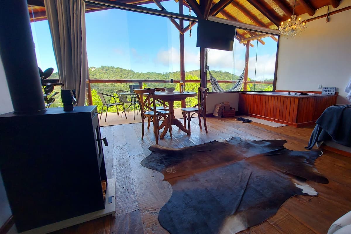 A room with a wooden floor, a dining table with chairs, a large window with a view of green hills, a bed with a blanket, a TV, and a guitar on the wall.