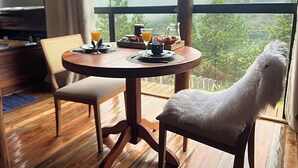 A dining area with a round wooden table, two chairs, and a view of a forest through large windows. - Chalés Luz do Luar (Campos do Jordao)