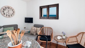 Una stanza con un tavolo rotondo, sedie in vimini, un divano, una sveglia a muro, una televisione e una finestra vista mare.