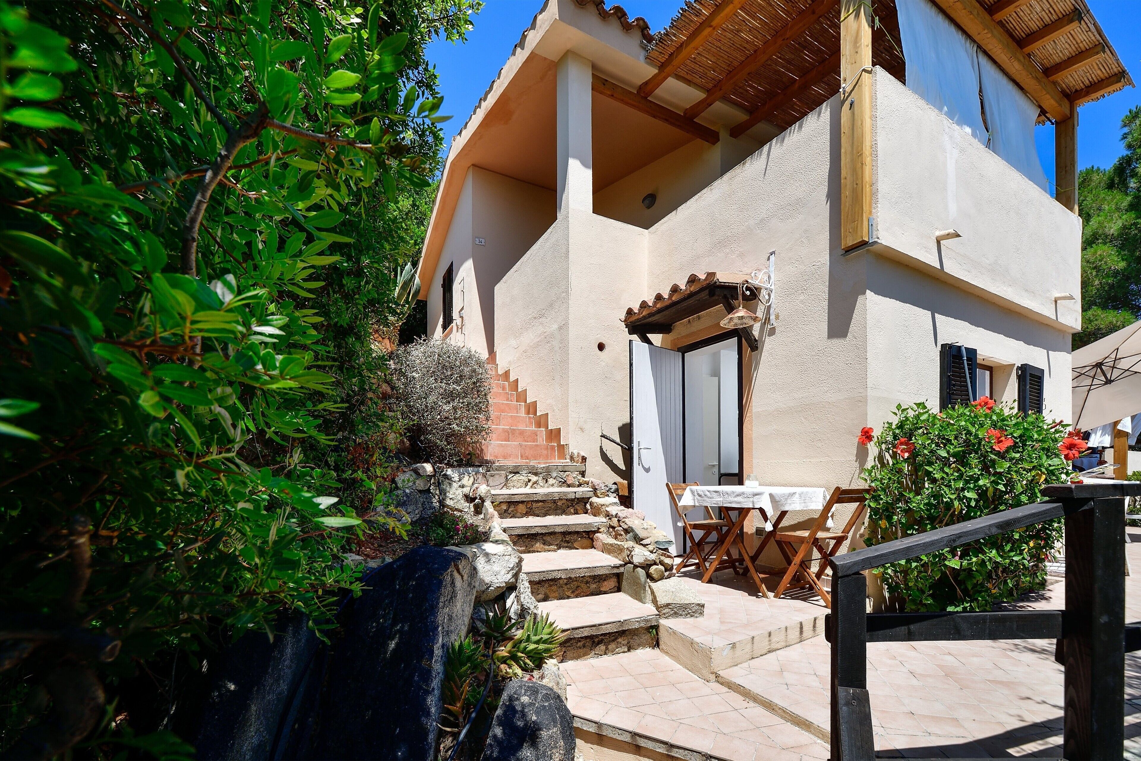 A house with a stone staircase leading to a door, a patio with a table and chairs, and a garden with various plants.