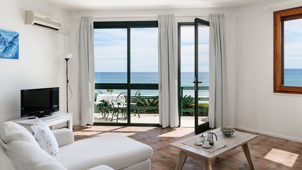 A modern living room with a television, a white sofa, a wooden coffee table, and a view of the sea through large glass doors. - Villa 'Massidda' with Sea View, Wi-Fi, and Air Conditioning (S.Margherita di Pula)