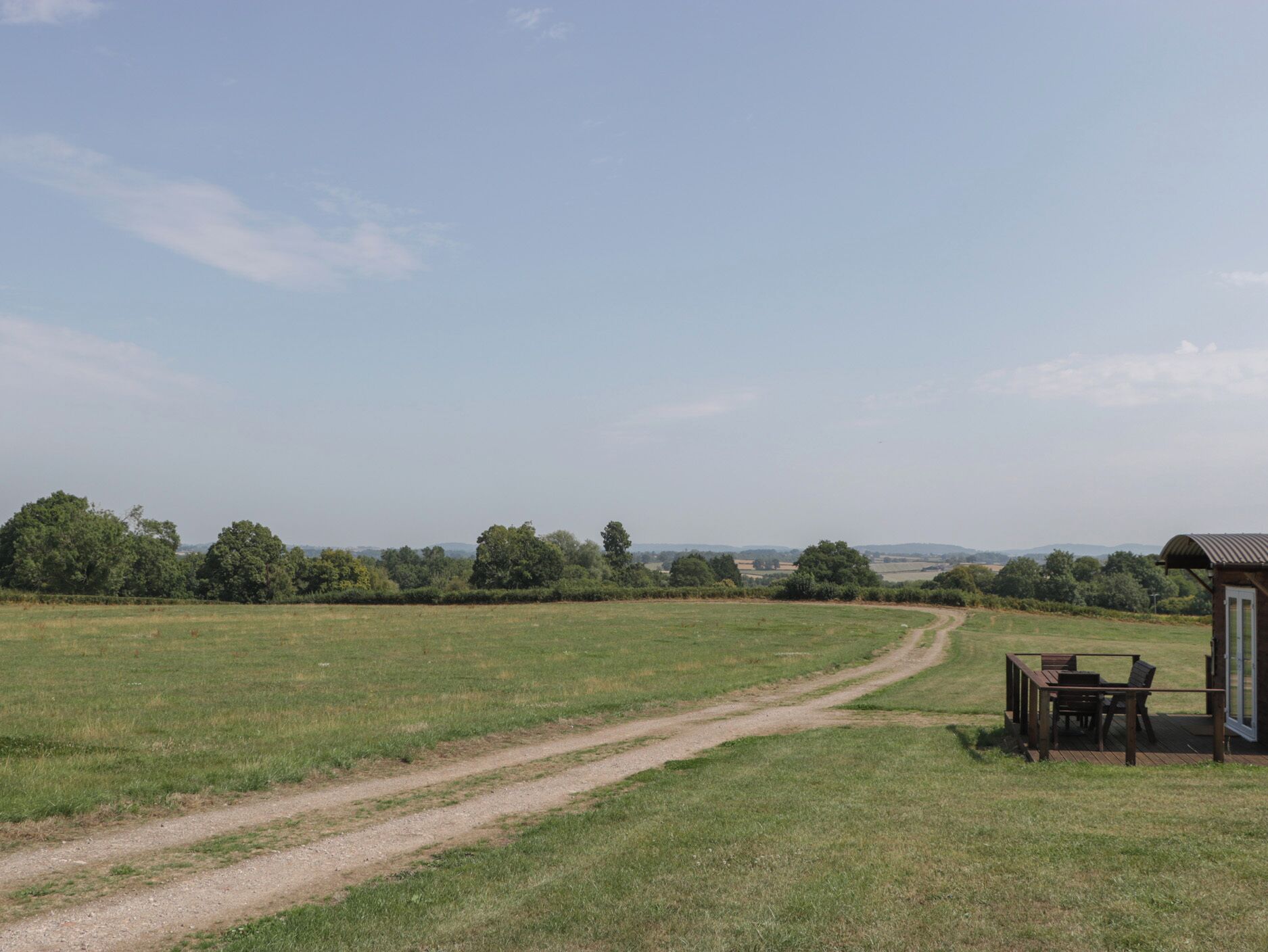 A rural landscape with a dirt path leading to a wooden structure with a table and chairs.