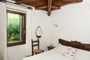 A bedroom with a wooden headboard, a bed with white bedding, a chair, a small table with a lamp, a window with curtains, and a wall-mounted light fixture.