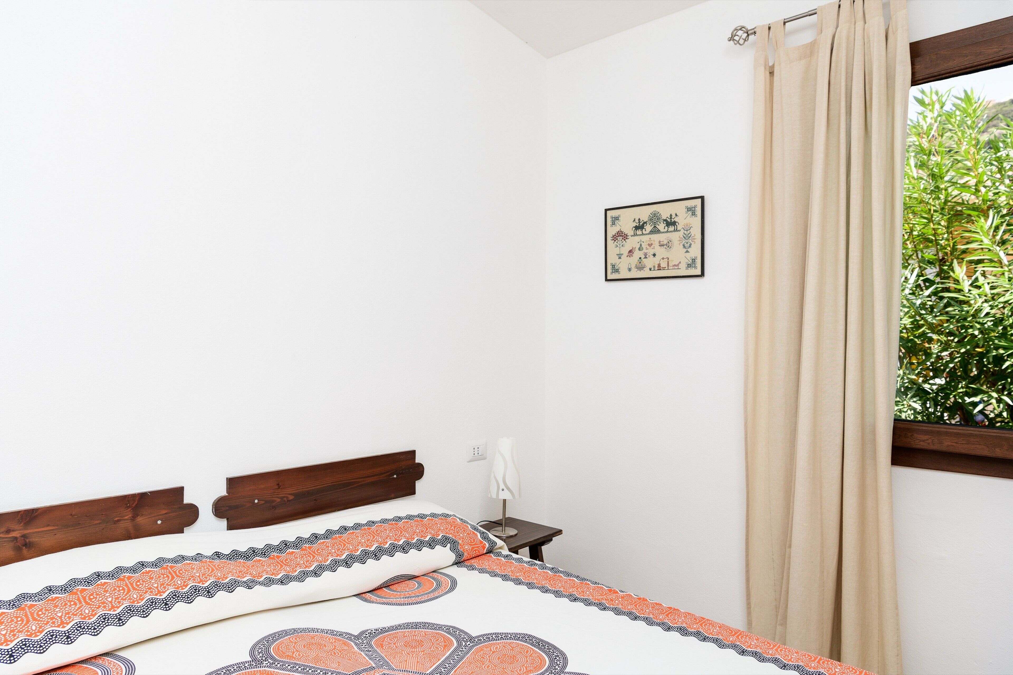 Ein Schlafzimmer mit Bett, Nachttisch, Wandlampe, Fenster mit Vorhängen und einem gerahmten Bild an der Wand.