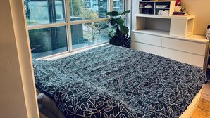 A bedroom with a large window, a bed with a leaf-patterned bedspread, a plant, and a white shelving unit with books and decorative items.