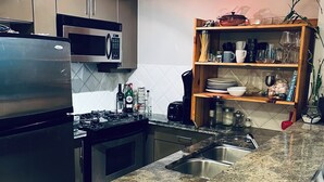A modern kitchen with stainless steel appliances, a granite countertop, and a wooden shelf with dishes and glasses.