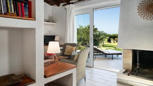 A room with a fireplace, a sofa, a table, a bookshelf, and a view of a garden through a glass door. - Villa 'Villetta 3' with Sea View, Wi-Fi and Air Conditioning (Costa Rei)