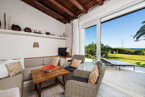A room with a sofa, a table, wicker chairs, a television, and a view of a green landscape through a sliding glass door.