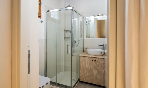 Un baño moderno con ducha envuelta en vidrio, tocador de madera y lavamanos blanco.